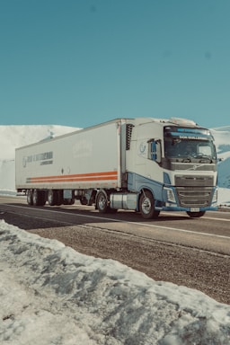 A large truck is driving along a snowy mountain road. The truck is mostly white with blue and orange detailing and features a long trailer. Snow-covered mountains and a clear blue sky dominate the background, creating a serene and calm landscape.