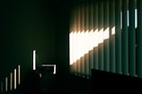 Vertical blinds partially opened on a window allow sunlight to enter a dimly lit room. The sunlight casts sharp, linear shadows on the opposite wall and small chest of drawers, highlighting the texture and creating a contrast between light and shadow.