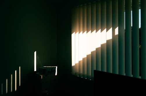 Vertical blinds partially opened on a window allow sunlight to enter a dimly lit room. The sunlight casts sharp, linear shadows on the opposite wall and small chest of drawers, highlighting the texture and creating a contrast between light and shadow.