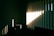Vertical blinds partially opened on a window allow sunlight to enter a dimly lit room. The sunlight casts sharp, linear shadows on the opposite wall and small chest of drawers, highlighting the texture and creating a contrast between light and shadow.