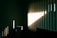 Vertical blinds partially opened on a window allow sunlight to enter a dimly lit room. The sunlight casts sharp, linear shadows on the opposite wall and small chest of drawers, highlighting the texture and creating a contrast between light and shadow.