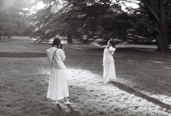 Two people dressed in long, white clothing are captured in a serene park setting surrounded by large trees and sunlight filtering through the branches. One person is taking a photograph of the other, who is standing with a contemplative pose.