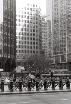 A monochrome urban scene featuring a row of bicycles parked at a bike-sharing station. Several individuals are interacting with the bicycles, and towering modern office buildings create a backdrop, with reflections visible on their glass facades. The street in the foreground adds to the cityscape setting.