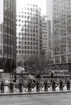 A monochrome urban scene featuring a row of bicycles parked at a bike-sharing station. Several individuals are interacting with the bicycles, and towering modern office buildings create a backdrop, with reflections visible on their glass facades. The street in the foreground adds to the cityscape setting.