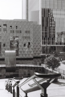 The image depicts a modern urban scene with large, geometric buildings featuring reflective glass surfaces. There is a unique, textured building facade and an architectural structure that seems to bridge over water. The scene includes railings leading down to the water and a solitary tree providing a touch of greenery.