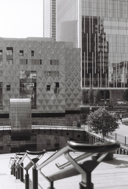 The image depicts a modern urban scene with large, geometric buildings featuring reflective glass surfaces. There is a unique, textured building facade and an architectural structure that seems to bridge over water. The scene includes railings leading down to the water and a solitary tree providing a touch of greenery.