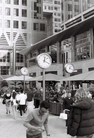 A bustling urban scene features several people walking in a public space surrounded by tall modern buildings. Large clocks are prominently displayed on poles, indicating time in front of a café or open-area restaurant with outdoor seating. The atmosphere is lively with a mix of individuals going about their daily activities.
