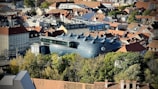 An aerial view of a cityscape featuring a large, modern art building with a unique, alien-like design and multiple round protrusions on the roof. The building is surrounded by traditional European architecture with red-tiled roofs and various trees.