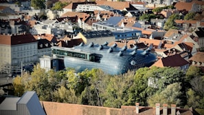 An aerial view of a cityscape featuring a large, modern art building with a unique, alien-like design and multiple round protrusions on the roof. The building is surrounded by traditional European architecture with red-tiled roofs and various trees.