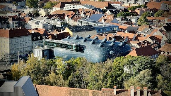 An aerial view of a cityscape featuring a large, modern art building with a unique, alien-like design and multiple round protrusions on the roof. The building is surrounded by traditional European architecture with red-tiled roofs and various trees.