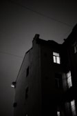 A dark, moody photograph of a building's corner at night, with light streaming from some of the windows. The sky appears cloudy, emphasizing the silhouette of the building's sharp angles and edges. Power lines stretch across the frame, adding to the urban atmosphere.