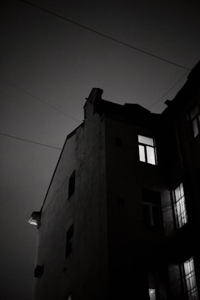 A dark, moody photograph of a building's corner at night, with light streaming from some of the windows. The sky appears cloudy, emphasizing the silhouette of the building's sharp angles and edges. Power lines stretch across the frame, adding to the urban atmosphere.