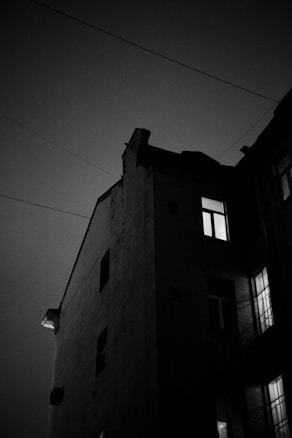 A dark, moody photograph of a building's corner at night, with light streaming from some of the windows. The sky appears cloudy, emphasizing the silhouette of the building's sharp angles and edges. Power lines stretch across the frame, adding to the urban atmosphere.