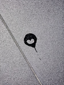 A dark liquid stain resembles the shape of a heart against a textured gray concrete surface. The stain is partially outlined by a lighter area, creating a contrast with the surrounding concrete.