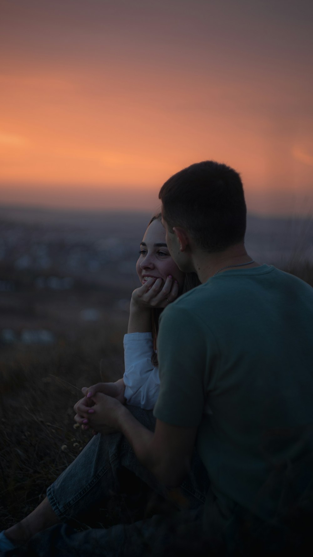 a man and a woman sitting on a hill at sunset