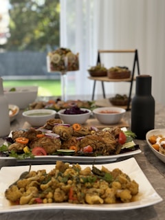 A dining table is set with various dishes including plates of fried and sautéed food, accompanied by bowls of salad and condiments. The scene is bright with natural lighting from the window, providing a homely and inviting atmosphere. The table is arranged aesthetically with a variety of foods showcasing vibrant colors.