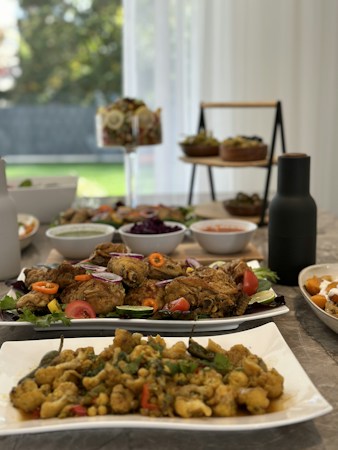 A dining table is set with various dishes including plates of fried and saut&eacute;ed food, accompanied by bowls of salad and condiments. The scene is bright with natural lighting from the window, providing a homely and inviting atmosphere. The table is arranged aesthetically with a variety of foods showcasing vibrant colors.