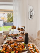 A dining table is set with a variety of colorful dishes, including fried chicken garnished with peppers and lime, dipping sauces in bowls, a fruit dessert in a glass container, and an array of appetizers arranged on a tiered stand. The table is located near a bright window with sheer white curtains, overlooking a green backyard with autumn trees.