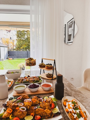 A dining table is set with a variety of colorful dishes, including fried chicken garnished with peppers and lime, dipping sauces in bowls, a fruit dessert in a glass container, and an array of appetizers arranged on a tiered stand. The table is located near a bright window with sheer white curtains, overlooking a green backyard with autumn trees.