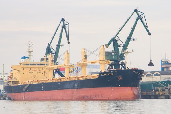 A large cargo ship named Harvester is docked at a port. It is equipped with cranes used for loading and unloading, with one crane currently lifting a load. The ship has a prominent 'No Smoking Safety First' sign displayed on its side. The backdrop includes a coastal industrial area with buildings and storage facilities.