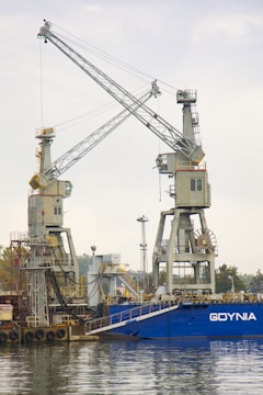 Two large industrial cranes are situated at a harbor with a blue ship docked nearby. The cranes are positioned over the water, and there is industrial machinery and infrastructure around them. The background shows a cloudy sky with some greenery and additional structures in the distance.