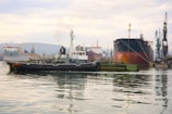 A busy industrial port with several ships docked at a wharf. A tugboat is in the foreground, pulling or guiding a massive cargo ship. Cranes and other port infrastructure are visible in the background, with reflections shimmering on the calm water.