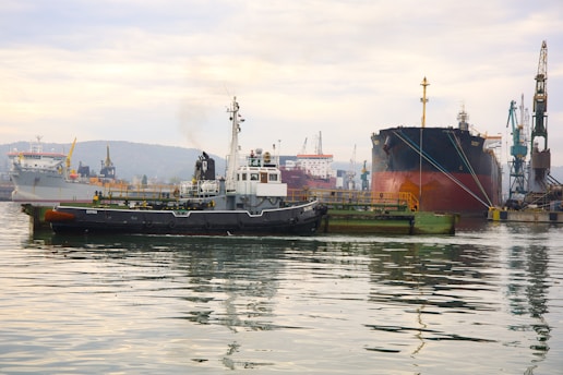 A busy industrial port with several ships docked at a wharf. A tugboat is in the foreground, pulling or guiding a massive cargo ship. Cranes and other port infrastructure are visible in the background, with reflections shimmering on the calm water.