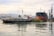 A busy industrial port with several ships docked at a wharf. A tugboat is in the foreground, pulling or guiding a massive cargo ship. Cranes and other port infrastructure are visible in the background, with reflections shimmering on the calm water.