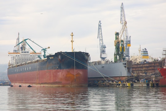 Several large cargo ships are docked at a bustling shipyard. Cranes and industrial equipment are visible, indicating active maintenance or loading operations. The water in the foreground reflects the cloudy sky above.