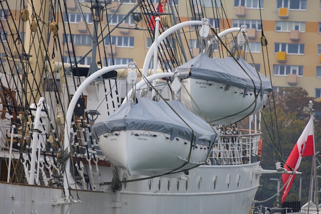 Two lifeboats are suspended on the side of a tall ship, with their names 'Dar Pomorza Gdynia' visible. The ship features a complex array of ropes and pulleys. In the background, a beige residential building with multiple windows is seen, along with a Polish flag.