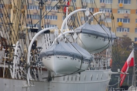Two lifeboats are suspended on the side of a tall ship, with their names 'Dar Pomorza Gdynia' visible. The ship features a complex array of ropes and pulleys. In the background, a beige residential building with multiple windows is seen, along with a Polish flag.