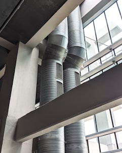 Two large, industrial air ducts with a metallic finish extend vertically alongside a building's structural beams. Natural light filters in from the expansive glass windows, which create geometric patterns with their framework. The scene is characterized by a modern, industrial aesthetic.