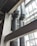 Two large, industrial air ducts with a metallic finish extend vertically alongside a building's structural beams. Natural light filters in from the expansive glass windows, which create geometric patterns with their framework. The scene is characterized by a modern, industrial aesthetic.