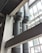 Two large, industrial air ducts with a metallic finish extend vertically alongside a building's structural beams. Natural light filters in from the expansive glass windows, which create geometric patterns with their framework. The scene is characterized by a modern, industrial aesthetic.