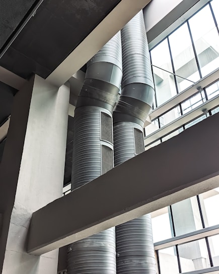 Two large, industrial air ducts with a metallic finish extend vertically alongside a building's structural beams. Natural light filters in from the expansive glass windows, which create geometric patterns with their framework. The scene is characterized by a modern, industrial aesthetic.