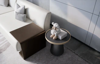 A modern living space features a light-colored sofa with geometric-patterned cushions. A small wooden side table is adjacent to the sofa, and a round side table holds a sitting statue in light tones, possibly resembling a character or figure.