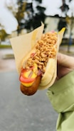 A hand holds a hot dog in a bun with crispy fried onions on top, along with ketchup and mustard drizzles. The hot dog is wrapped partially in a paper sleeve. The background shows an outdoor setting with blurred trees and buildings.