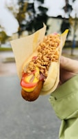 A hand holds a hot dog in a bun with crispy fried onions on top, along with ketchup and mustard drizzles. The hot dog is wrapped partially in a paper sleeve. The background shows an outdoor setting with blurred trees and buildings.