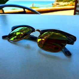 A pair of sunglasses with reflective lenses sits on a white surface, capturing the scenic view of a beach in the background. The lenses reflect the blue sky and some greenery. The table seems to be located outdoors, near a coastal area with a railing visible in the background.