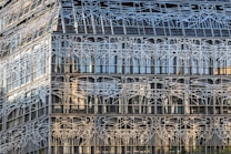 A modern building facade with intricate, abstract metal latticework covering large glass panels. The design consists of numerous intersecting lines creating an organic, web-like pattern.