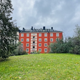 A red brick apartment building with multiple windows stands prominently against a cloudy sky. Surrounding the building are lush green trees and a grassy lawn with scattered autumn leaves.