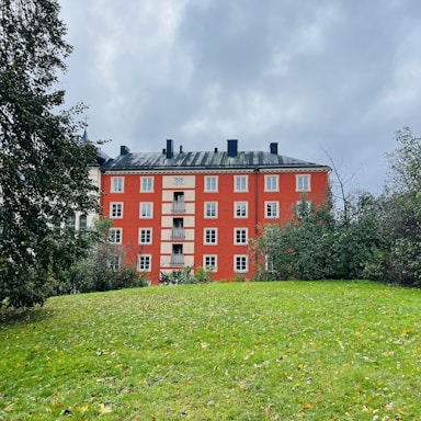 A red brick apartment building with multiple windows stands prominently against a cloudy sky. Surrounding the building are lush green trees and a grassy lawn with scattered autumn leaves.