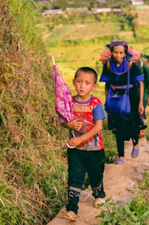 A young boy is walking along a narrow path beside lush green vegetation, holding a pink patterned umbrella. Behind him, an older woman is following, wearing traditional attire with bright blue accents. The background shows cultivated fields and distant foliage, suggesting a rural setting.