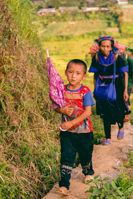 A young boy is walking along a narrow path beside lush green vegetation, holding a pink patterned umbrella. Behind him, an older woman is following, wearing traditional attire with bright blue accents. The background shows cultivated fields and distant foliage, suggesting a rural setting.
