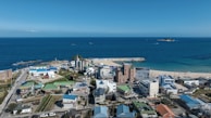 A coastal town with a mix of low-rise residential and commercial buildings, featuring a small beach area and a pier extending into the ocean. The landscape includes a mix of green areas and paved roads, with a vast blue sea and clear sky meeting at the horizon.