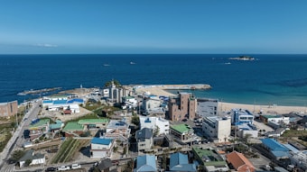 A coastal town with a mix of low-rise residential and commercial buildings, featuring a small beach area and a pier extending into the ocean. The landscape includes a mix of green areas and paved roads, with a vast blue sea and clear sky meeting at the horizon.