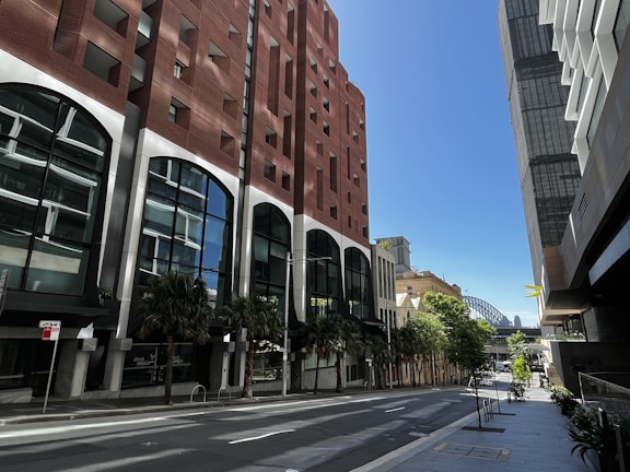 A city street lined with tall buildings, including a brick structure with large windows on one side and a modern glass building on the other. Trees are interspersed along the sidewalk, which appears empty of people and vehicles. In the background, a recognizable steel bridge arches over the horizon under a clear blue sky.