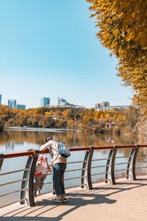 Two people, possibly a parent and child, are leaning over a railing by a calm lake, surrounded by trees with autumn foliage. In the background, there are modern buildings that dot the skyline under a clear blue sky.