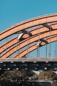 An industrial-style bridge with large, curved orange steel beams is set against a clear blue sky. The bridge features vertical and diagonal support structures and streetlights can be seen along the top. In the background, trees and urban structures are partially visible underneath.