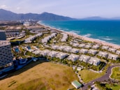 The scene captures a coastal resort area with numerous white buildings arranged in neat rows parallel to the coastline. The buildings are surrounded by lush greenery and well-maintained gardens. The turquoise ocean stretches alongside the sandy beach, while mountains rise in the background under a clear blue sky.